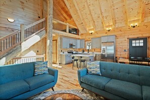 A spacious cabin interior with a high ceiling, wooden walls, a kitchen with modern appliances, a dining area, and a cozy seating area with blue sofas.