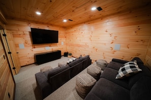 A wooden-paneled room with a flat-screen TV, dark sofas, and woven ottomans.