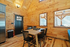 A wooden cabin interior with a dining table, chairs, a black door, and a window with a view of trees.