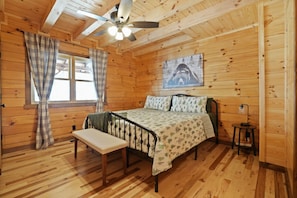 Un dormitorio de cabaña de madera con una cama, banca, ventilador de techo y una ventana con cortinas.