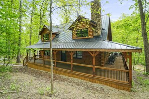 A rustic cabin with a wooden deck and a stone chimney, surrounded by a forest.