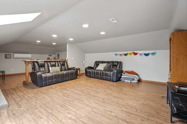 A spacious living room with a high ceiling, wooden flooring, two leather sofas, a small wooden table, and a wall-mounted air conditioner.