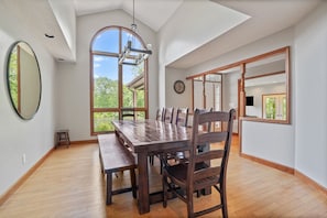 A dining area with a large wooden table and chairs, a hanging light fixture, and a view of greenery through a large window. - Deer Ridge Lodge | Hocking Hills, Sleeps 26, Pool (Laurelville)