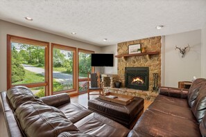 A spacious living room with a stone fireplace, leather sofas, and large windows offering a view of a road and greenery. - Deer Ridge Lodge | Hocking Hills, Sleeps 26, Pool (Laurelville)
