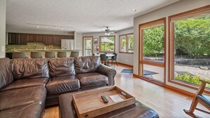 A spacious living area with a leather sofa, wooden coffee table, and large windows offering a view of the outdoors. - Deer Ridge Lodge | Hocking Hills, Sleeps 26, Pool (Laurelville)