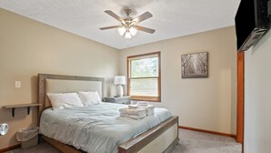 A bedroom with a bed, a nightstand, a lamp, a ceiling fan, a window with blinds, and a picture on the wall. - Deer Ridge Lodge | Hocking Hills, Sleeps 26, Pool (Laurelville)
