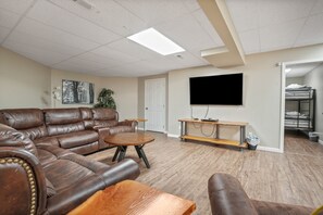 A living room with a brown leather sectional sofa, a wooden coffee table, a flat-screen TV mounted on the wall, and a small wooden side table. - Deer Ridge Lodge | Hocking Hills, Sleeps 26, Pool (Laurelville)