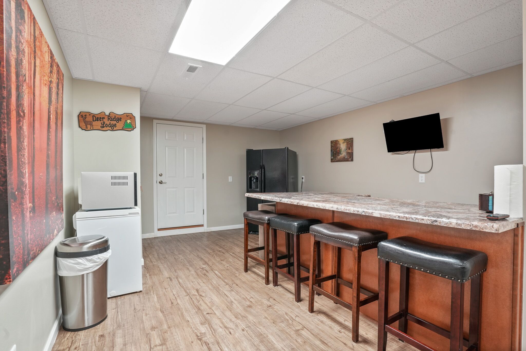A kitchen with a marble countertop, wooden stools, a refrigerator, a trash can, and a television mounted on the wall.
