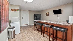 A kitchen with a marble countertop, wooden stools, a refrigerator, a trash can, and a television mounted on the wall. - Deer Ridge Lodge | Hocking Hills, Sleeps 26, Pool (Laurelville)