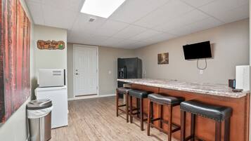 A kitchen with a marble countertop, wooden stools, a refrigerator, a trash can, and a television mounted on the wall.