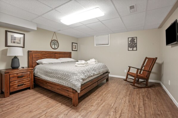 A neatly arranged bedroom with a wooden bed, a nightstand with drawers, a chair, and a television on the wall. - Deer Ridge Lodge | Hocking Hills, Sleeps 26, Pool (Laurelville)