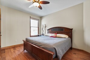 Une chambre avec un lit en bois, un ventilateur de plafond, une fenêtre avec des stores et une petite table avec une lampe.