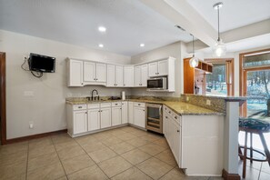 Cocina moderna con armarios blancos, encimeras de granito, horno integrado y un amplio ventanal con vistas a árboles.