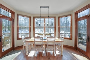 Un coin repas avec une table et des chaises en bois, de grandes fenêtres donnant sur une terrasse enneigée et un luminaire suspendu.