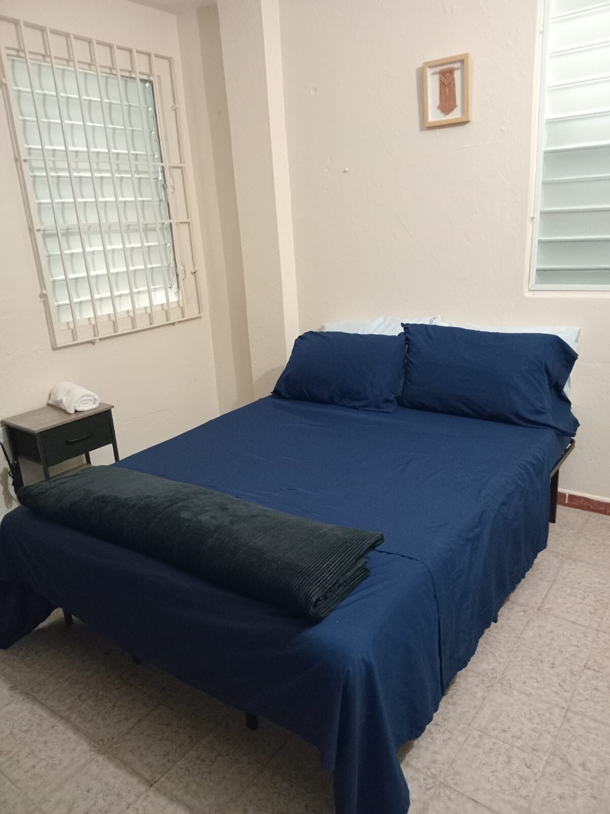 A bedroom with a bed, a nightstand, a window with bars, and a framed picture on the wall.