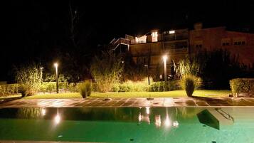 Uma área de piscina à noite, com piscina transparente, rodeada por caminhos bem iluminados e vegetação.