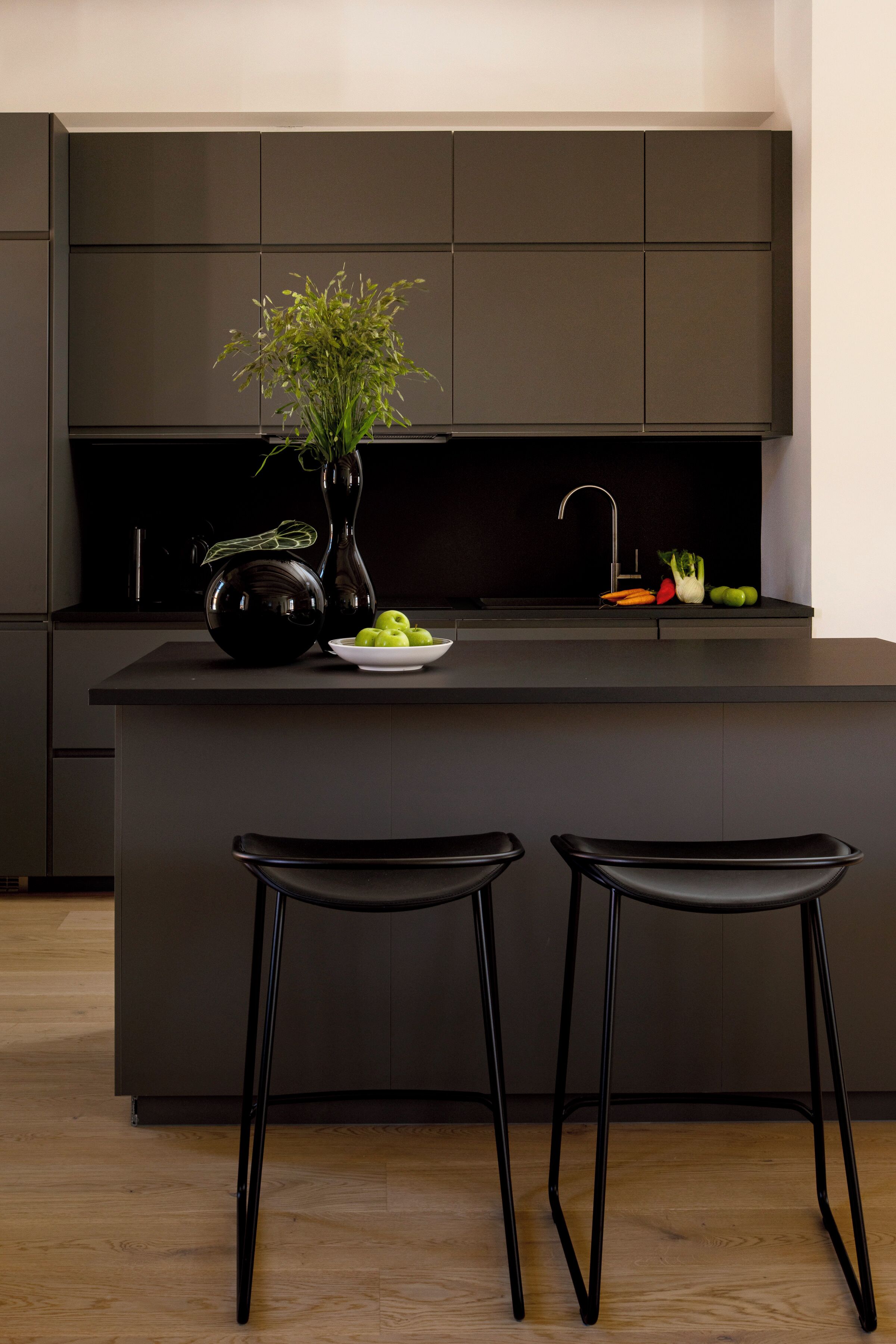 Une cuisine moderne avec un îlot central, deux tabourets de bar noirs, un vase noir contenant des plantes vertes et un saladier rempli de pommes vertes.
