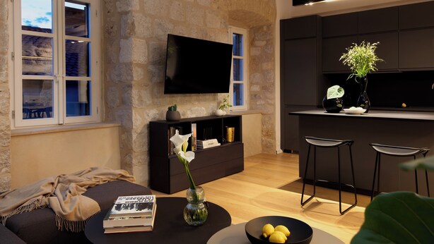A modern living room with a stone wall, a flat-screen TV, a sofa, a coffee table with a book and a vase, and a kitchen area with stools.
