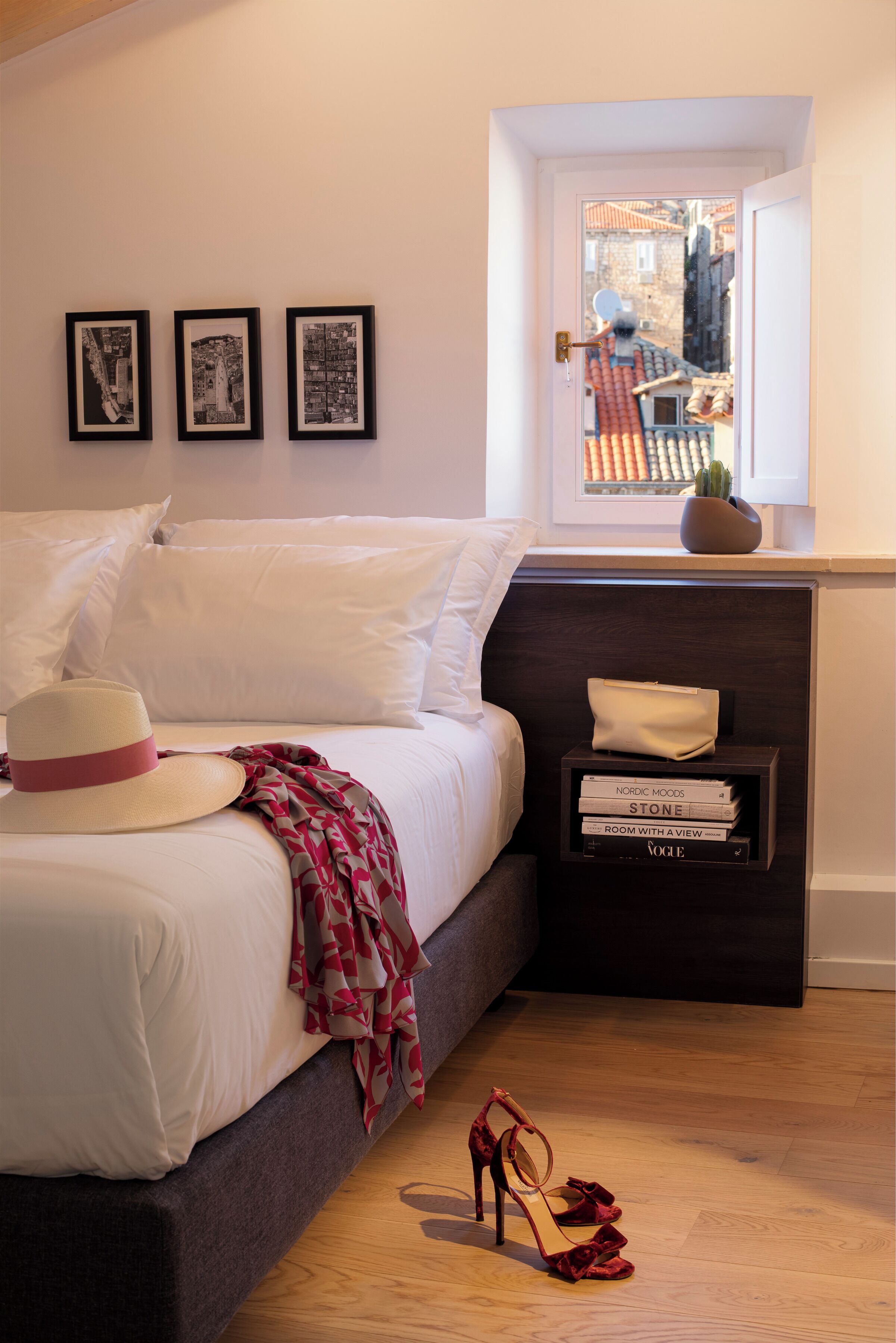 A neatly made bed with white linens, a red and white patterned throw, a beige hat, and a pair of red high heels on the floor. There are three framed black and white photographs on the wall above the bed. A small bedside table holds a few books and a white object.