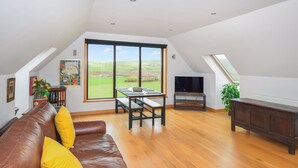 A spacious loft with a high ceiling, a large window offering a view of the countryside, a leather sofa with yellow cushions, a wooden dining table, and a television on a stand.