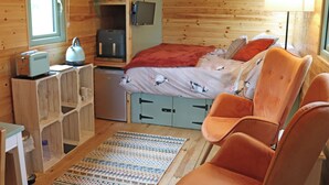 Ein gemütlicher Raum mit Bett, Schreibtisch mit Lampe, Stuhl und einer kleinen Küchenzeile.