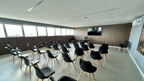 A conference room with rows of black chairs and tables, large windows, a flat-screen TV, and a wooden panel wall. - Edifício New Time - Apto1406 (Maceió)