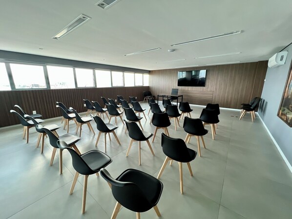 Un salón de conferencias con filas de sillas negras y mesas, ventanas amplias, un televisor de pantalla plana y un revestimiento de pared de madera.
