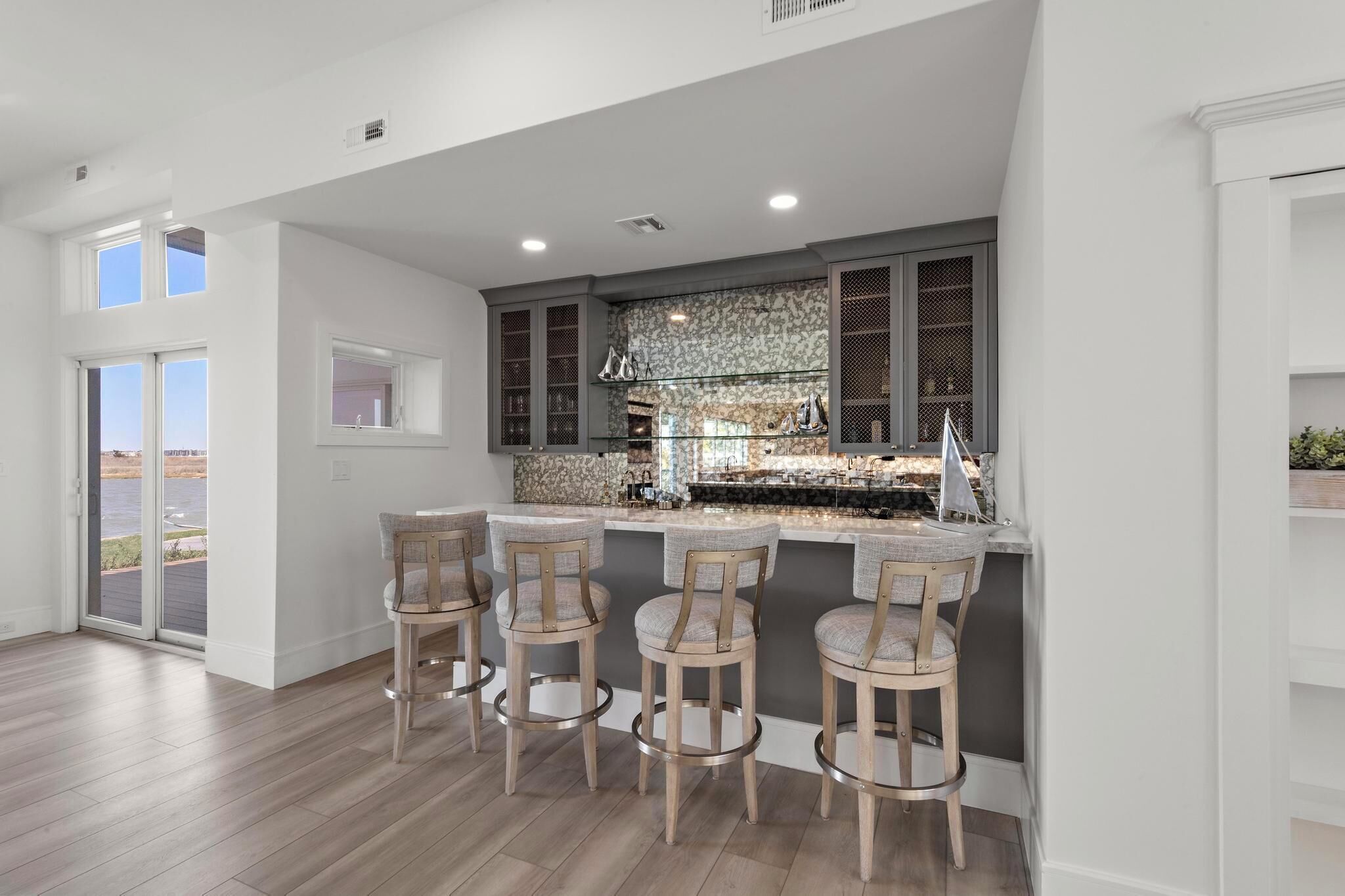 A modern kitchen with a bar area, featuring a stone backsplash, wooden bar stools, and a view through the glass doors.