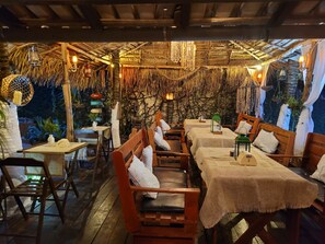 Un coin repas rustique avec des tables et des bancs en bois, un plafond recouvert de chaume et des lanternes suspendues.
