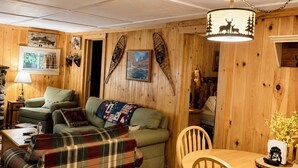 A cozy cabin interior with a plaid sofa, a green couch, a wooden dining table with chairs, and wooden walls. - Dog Friendly 🐾 Sleeps 8 • Near North Conway & Kancamangus Hwy. • Lake Access (Madison)