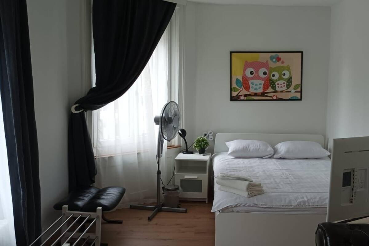 A bedroom with a bed, a desk, a chair, a standing fan, and a wall-mounted TV.