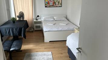 Ein kleiner, hell eingerichteter Raum mit einem Bett, einem Nachttisch und einem Schreibtisch mit Stuhl. Es gibt ein Fenster mit Vorhängen, einen kleinen Tisch mit einer Pflanze und einen Teppich auf dem Boden.