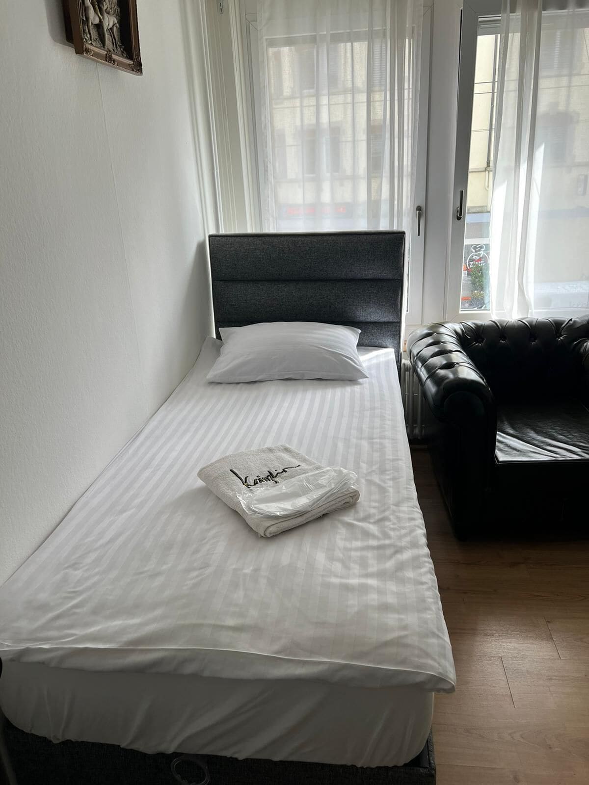 Ein Einzelbett mit grauem Kopfteil, weißer Bettwäsche und einem Handtuch mit einem Namen darauf. Eine schwarze Ledersofa befindet sich rechts.