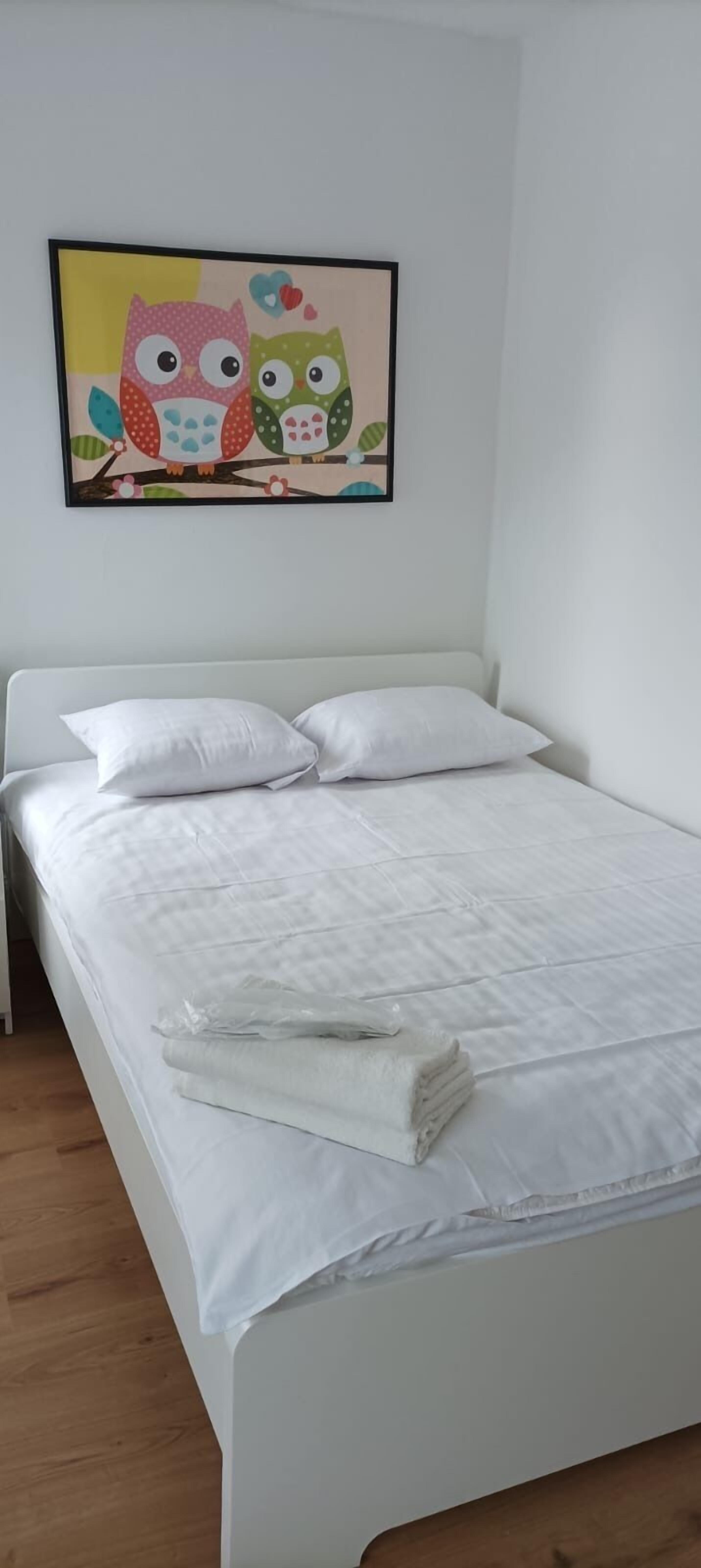 A neatly made bed with white bedding and pillows, a framed picture of owls on the wall, and a wooden floor.
