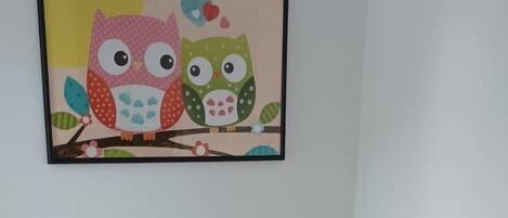 A neatly made bed with white bedding and pillows, a framed picture of owls on the wall, and a wooden floor.