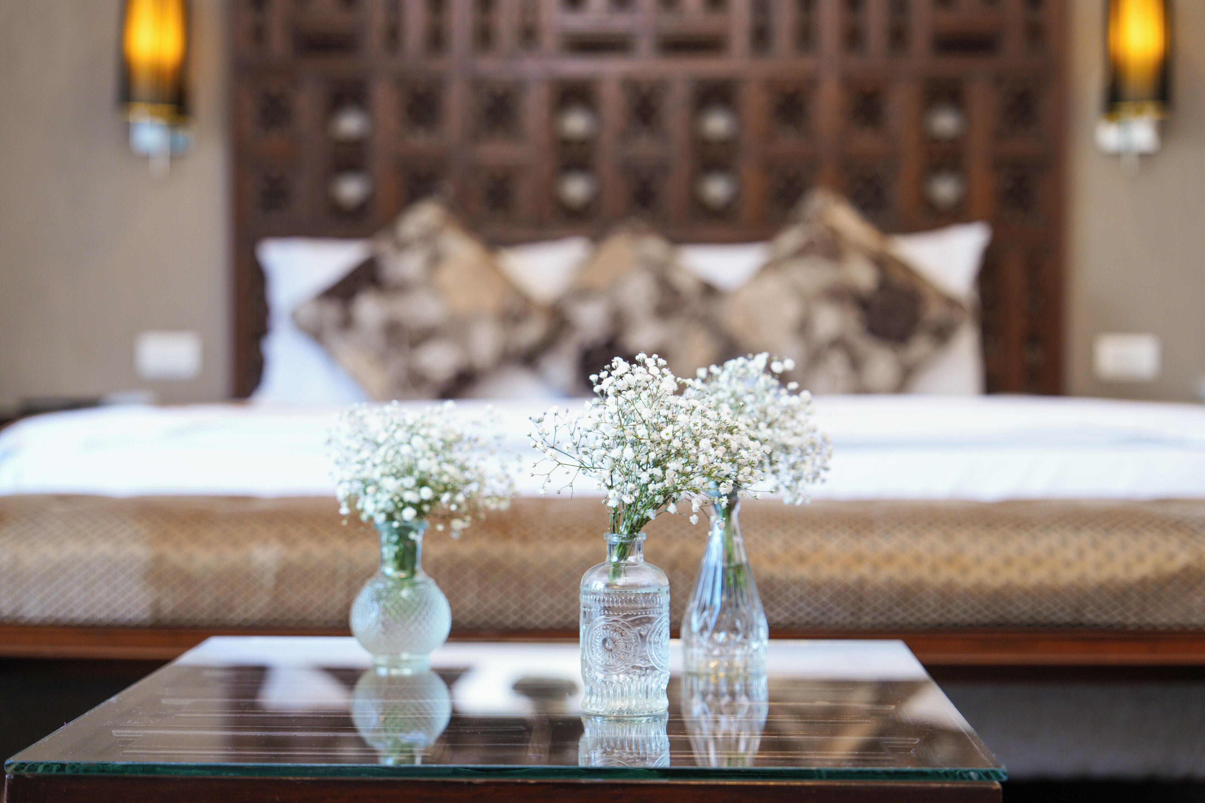 A bedroom with a bed, pillows, and two glass vases with flowers on a glass table.