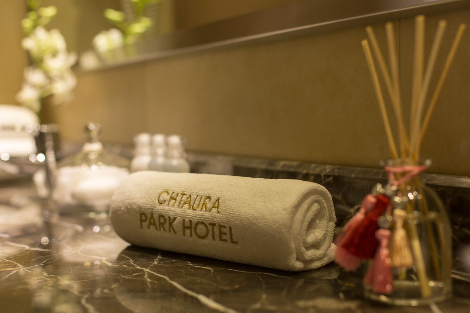 A rolled towel with "CHITAURA PARK HOTEL" embroidered on it, placed on a marble surface with a glass bottle and reed diffuser in the background.