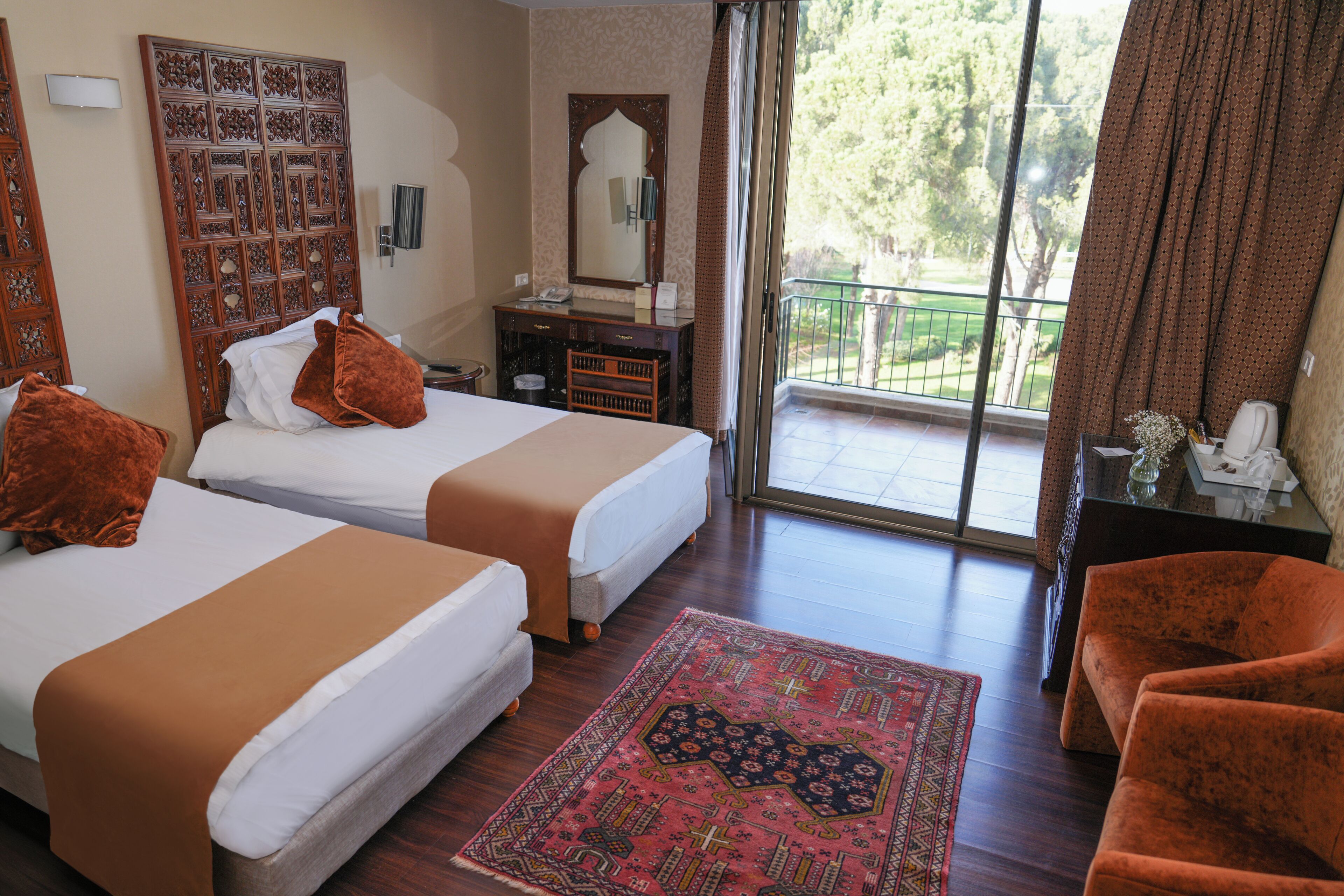 Habitación de hotel con dos camas, un balcón con vistas, cabecera de madera, una mesita con un florero y una alfombra estampada.