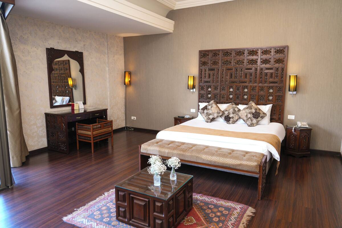 A spacious bedroom with a large bed, a wooden headboard, a nightstand, a dresser, a mirror, and a small table with a vase.