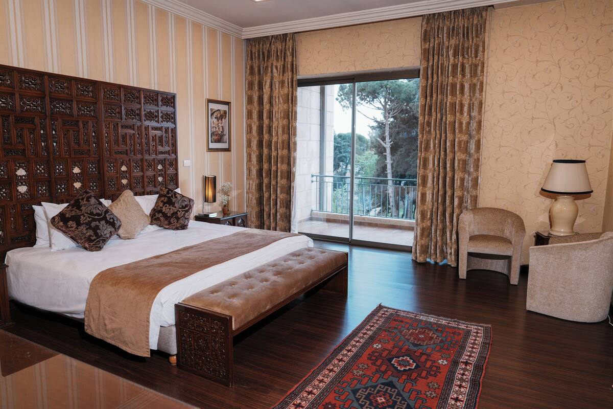 A spacious bedroom with a large bed, a wooden headboard, a balcony with curtains, and a view of trees.