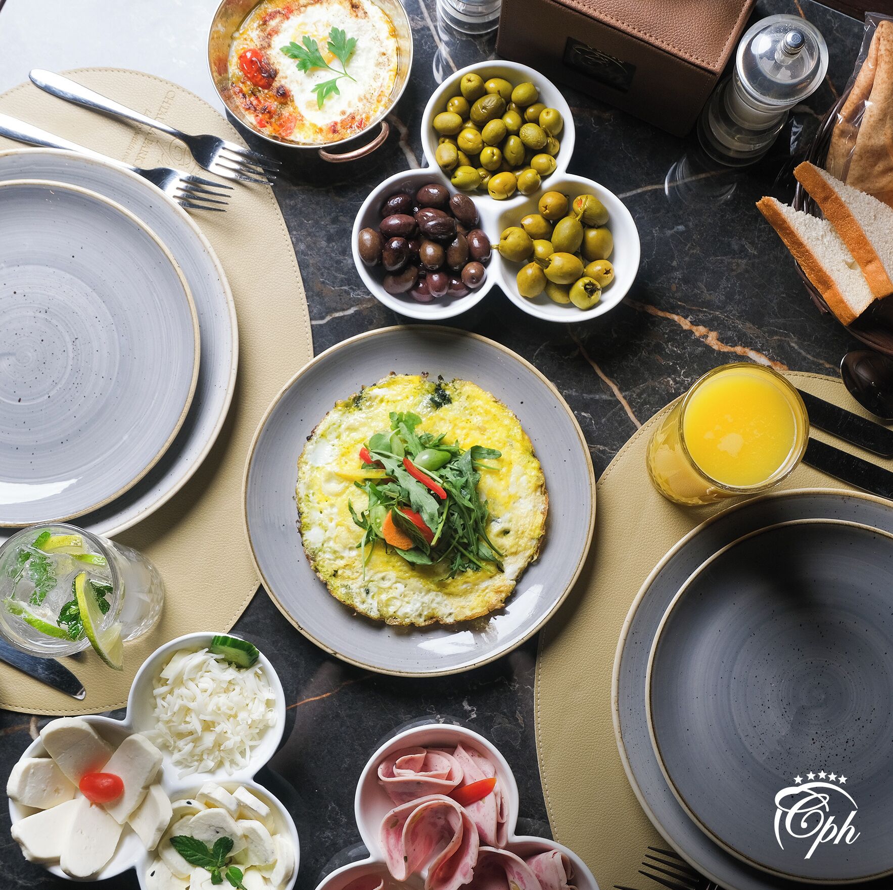 A breakfast setting with plates of food, olives, olives, and a glass of orange juice.