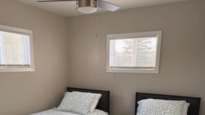 Quarto com duas camas de solteiro, ventilador de teto e duas janelas com persianas.