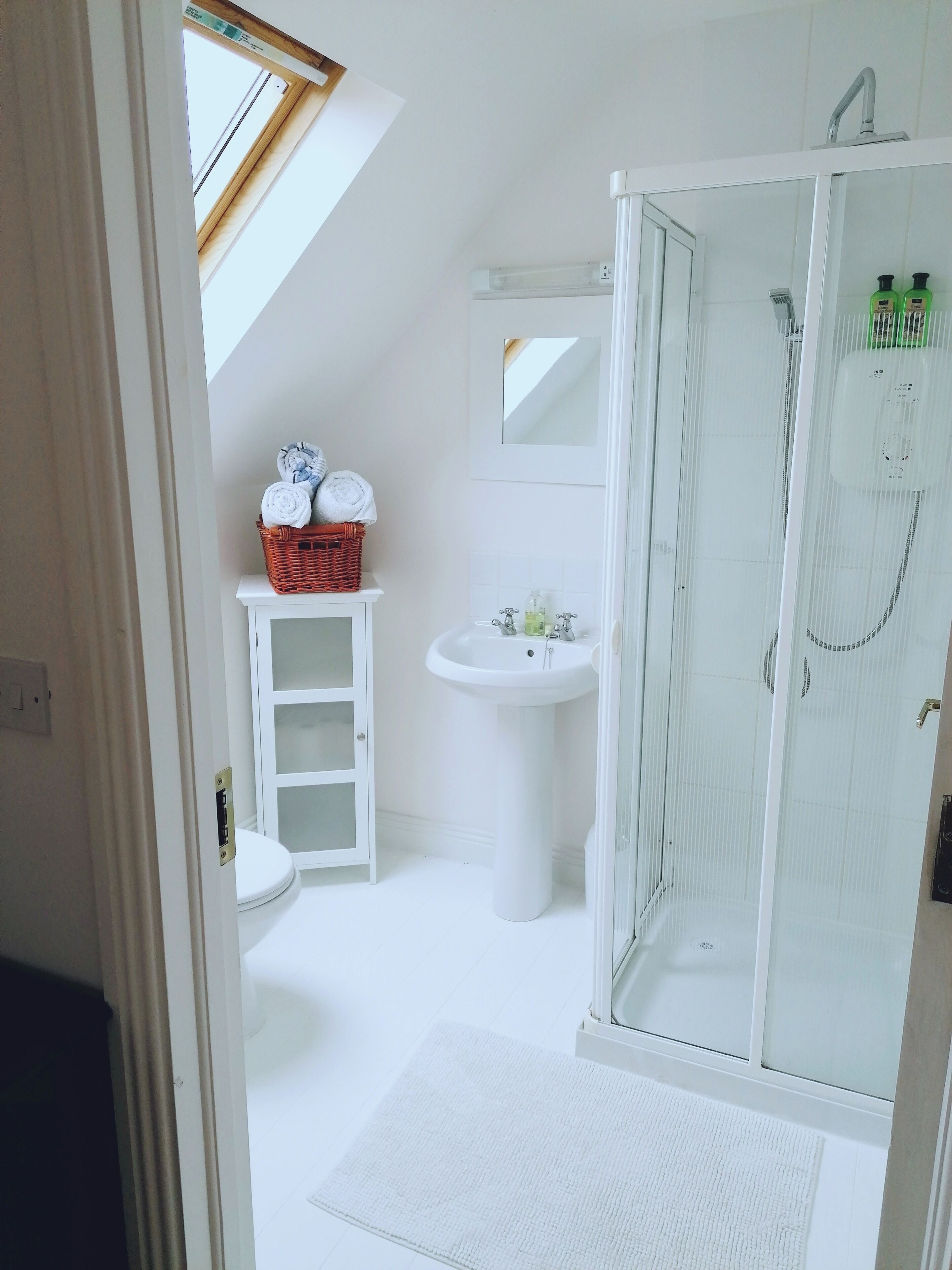 Ein Badezimmer mit Oberlicht, einem weißen Waschbecken, einer Duschkabine aus Glas und einem Korbwäscheschrank mit Handtüchern.