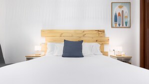 A neatly made bed with a wooden headboard, white bedding, and a single blue pillow. There are two bedside tables with lamps and a framed picture on the wall.