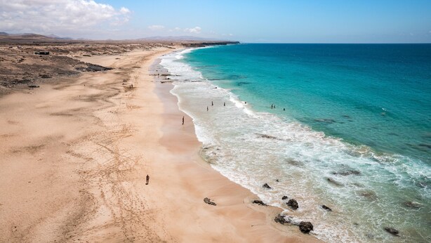 A wide sandy beach with clear turquoise waters and a few people walking along the shore.