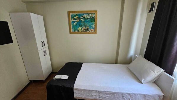 Un lit simple avec un matelas blanc et une couvre-lit noire, une armoire blanche à trois portes et un tableau représentant un étang fleuri accroché au mur.