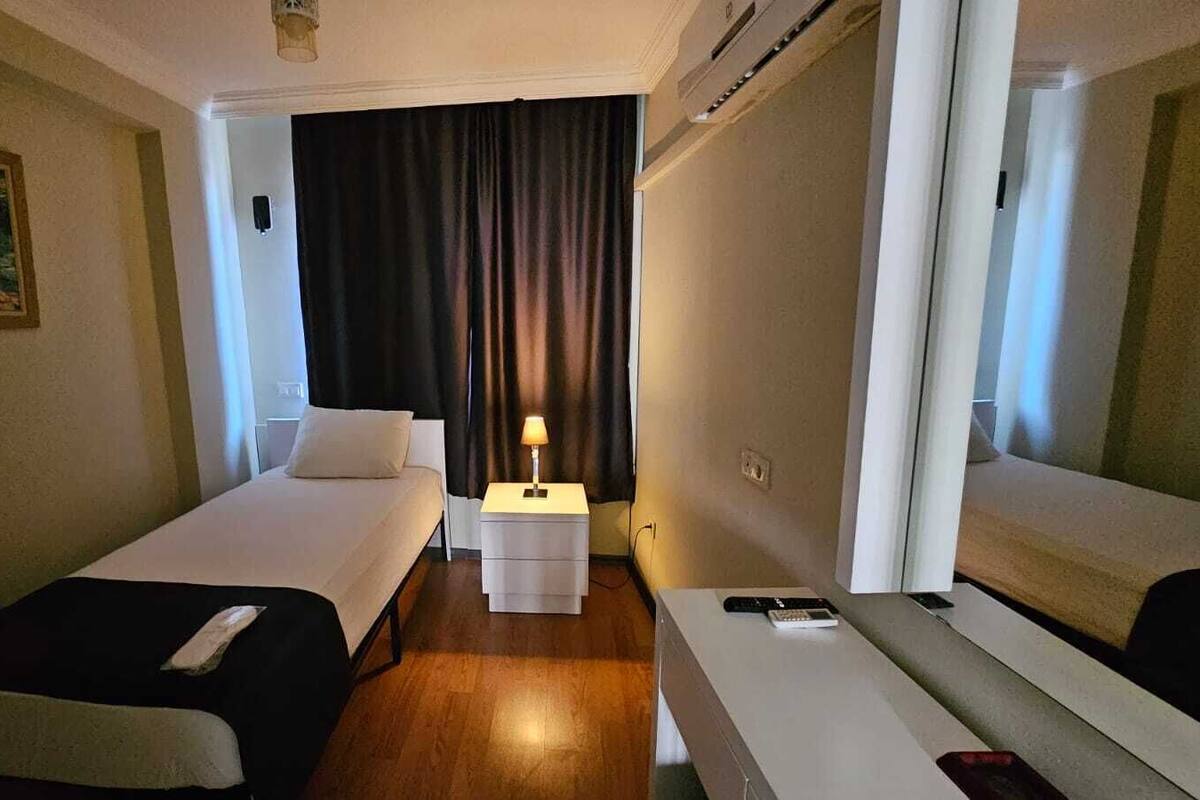Habitación de hotel con dos camas individuales, una mesita de noche con lámpara, un espejo grande y un control remoto sobre una mesita auxiliar.