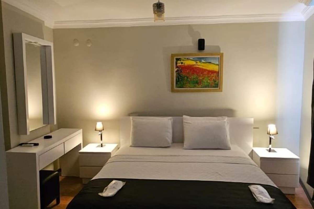 A hotel room with a bed, bedside tables, a desk, and a painting on the wall.
