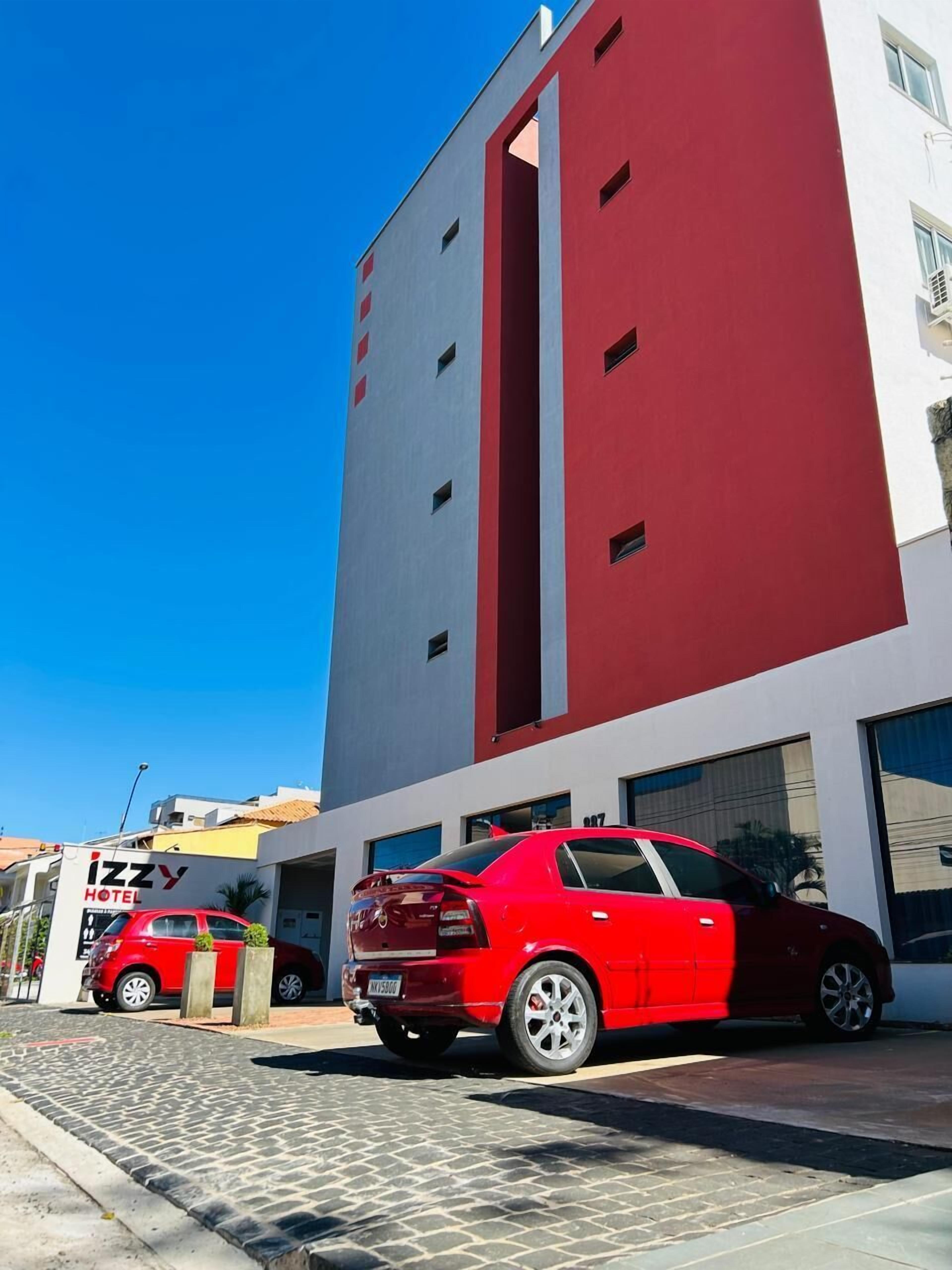 A modern building with a red and white facade, a red car parked in front, and a sign for 'IZZY HOTEL' visible.
