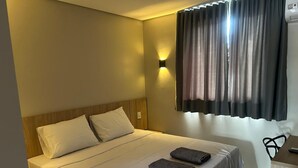 Een net opgemaakt bed met witte lakens en twee opgevouwen handdoeken, een nachtkastje met een afstandsbediening, en een wandlamp.